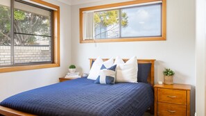 Un dormitorio con una cama grande, cabecera de madera, mesita de noche con una planta y ventana con vista a árboles.
