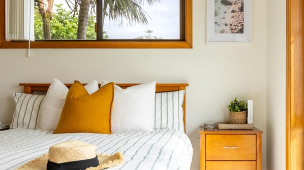 Un dormitorio ordenado con cama, almohadas, una mesita de noche con una planta y un cuadro en la pared.