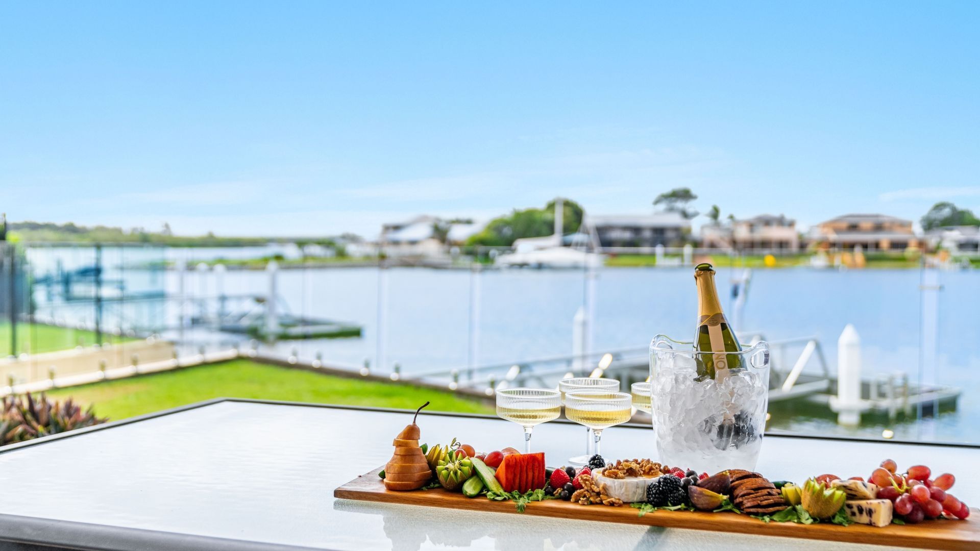 A table with a variety of fruits, nuts, and cheeses, a bottle of champagne, and two glasses of white wine, set against a backdrop of a waterfront view.