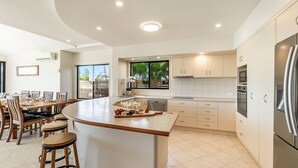 A modern kitchen with a central island, built-in appliances, and a dining area with wooden chairs. - Nirvana Waters - Absolute Waterfront Escape with Private Pontoon & Pool - Holiday Yamba (YAMBA)