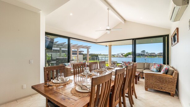 A dining area with a wooden table set for six, large windows offering a view of a body of water and buildings, and a ceiling fan. - Nirvana Waters - Absolute Waterfront Escape with Private Pontoon & Pool - Holiday Yamba (YAMBA)