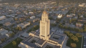 An aerial view of a large, historic building with a central tower and a dome, surrounded by parking and green spaces.