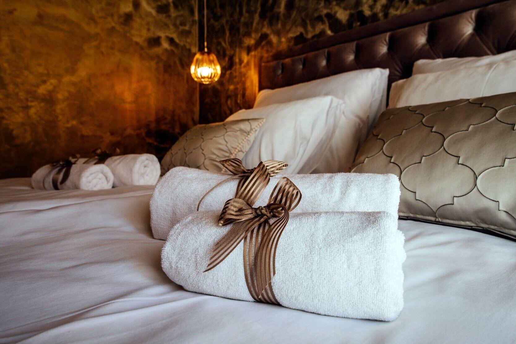 A neatly made bed with rolled towels and a decorative ribbon, a tufted headboard, and a hanging light fixture.