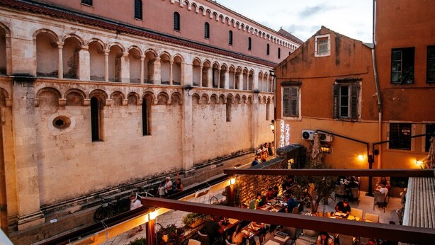 An outdoor dining area with tables and chairs, a stone building with arches, and a staircase leading to an upper level. - Luxury Rooms VINDEMIÆ (Zadar)