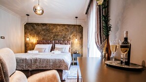 A hotel room with a large bed, a bedside table with a bottle of wine and glasses, a chair, and a view of the city. - Luxury Rooms VINDEMIÆ (Zadar)