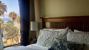 Quarto de hotel com cama, mesa de cabeceira, luminária e vista para palmeiras e a praia.
