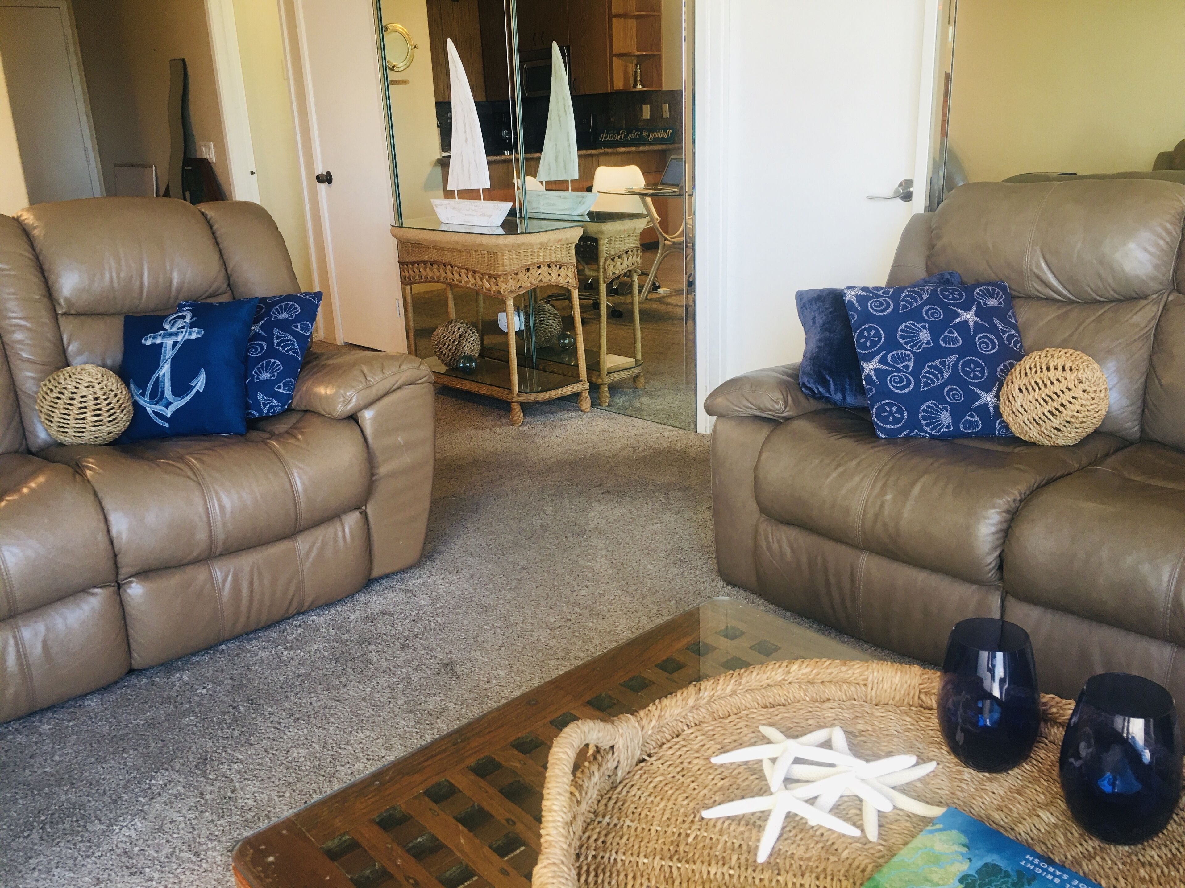 Een woonkamer met twee bruine leren banken, elk met een blauw kussen met een nautisch ankerdesign. In het midden staat een rieten salontafel met zeesterdecoraties.