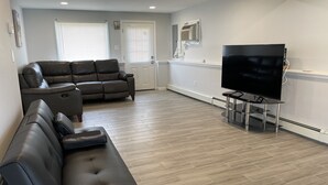 Una sala moderna con un sofá seccional, un televisor de pantalla plana sobre un mueble bajo y un gran ventanal con persianas.