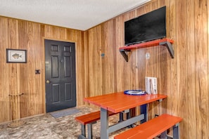 A room with wooden panel walls, a black door, a TV mounted on a red shelf, a table with benches, and a framed picture on the wall.