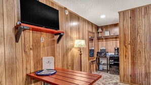 Una habitación con paneles de madera, un televisor montado en la pared, una mesa con una bandeja de revistas, una lámpara y una cafetera.