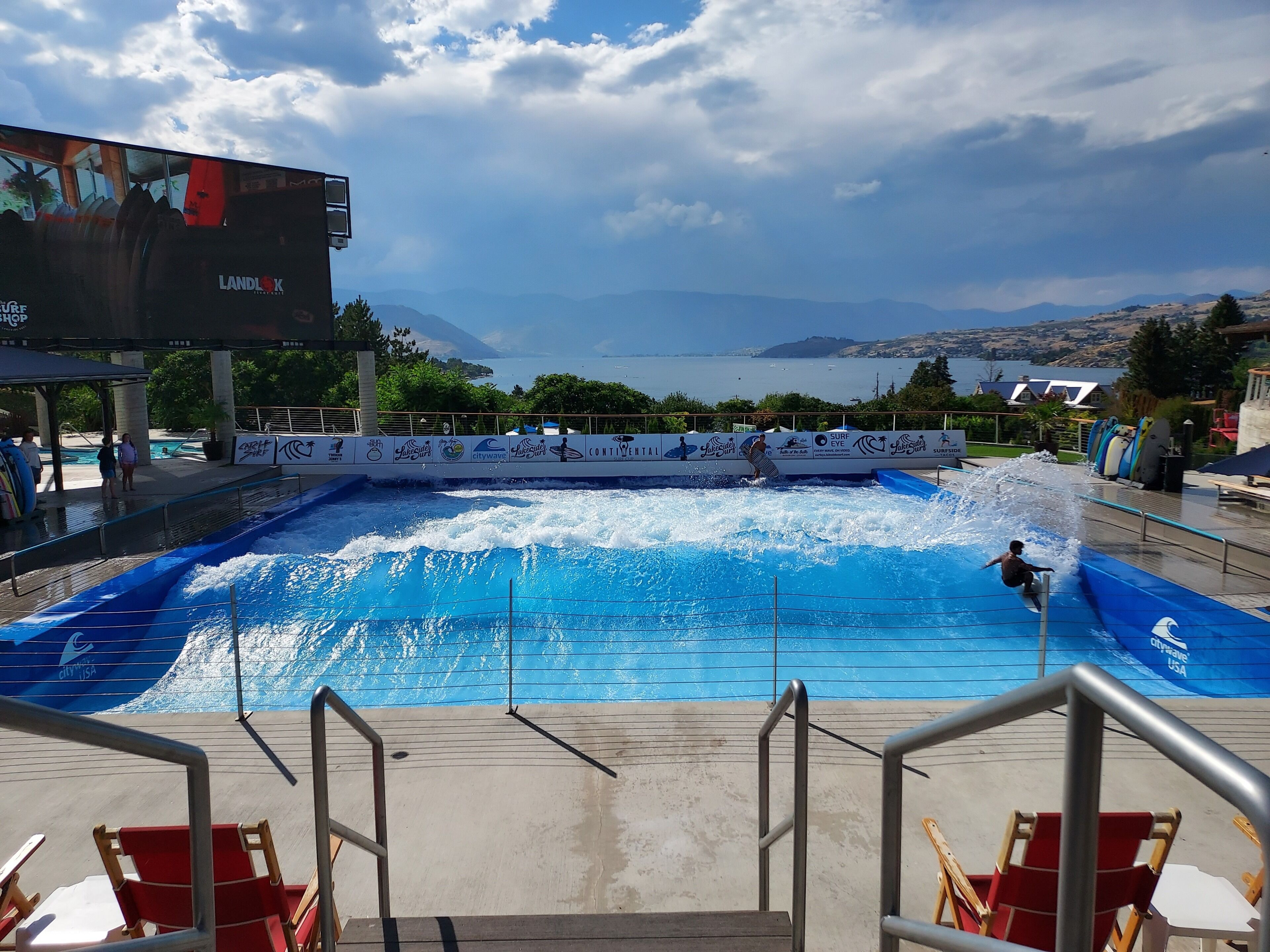 Une piscine avec une machine à vagues, entourée d’une terrasse dotée de chaises et offrant une vue sur la mer et les montagnes.