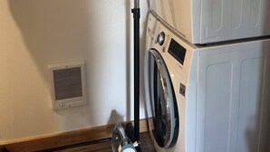 Un espace buanderie équipé d’une pile de machines à laver et de sèche-linge, d’une bouche d’aération et d’une étagère en métal.