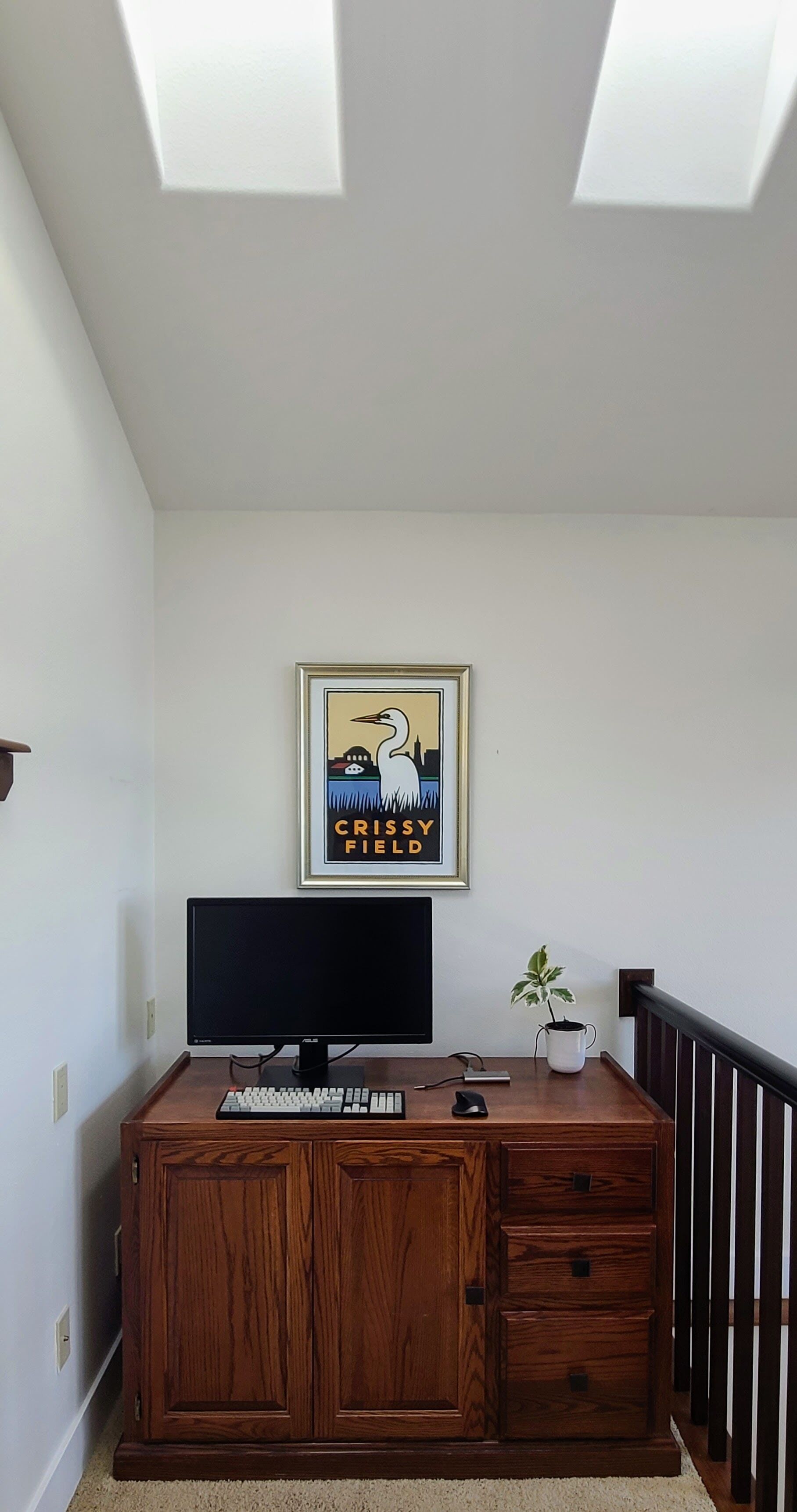 A room with a wooden desk, a computer monitor, a keyboard, a mouse, and a framed picture on the wall.