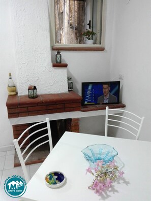 Ein Zimmer mit weißer Wand, einem Kamin aus Backsteinen, einem Fernseher und einem weißen Tisch mit Stühlen.