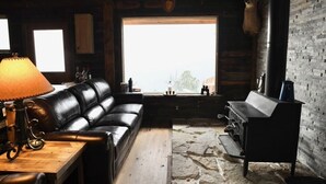 L’intérieur d’un chalet rustique, avec un canapé en cuir, une table avec une lampe, un sol en pierre, une grande fenêtre et un barbecue.