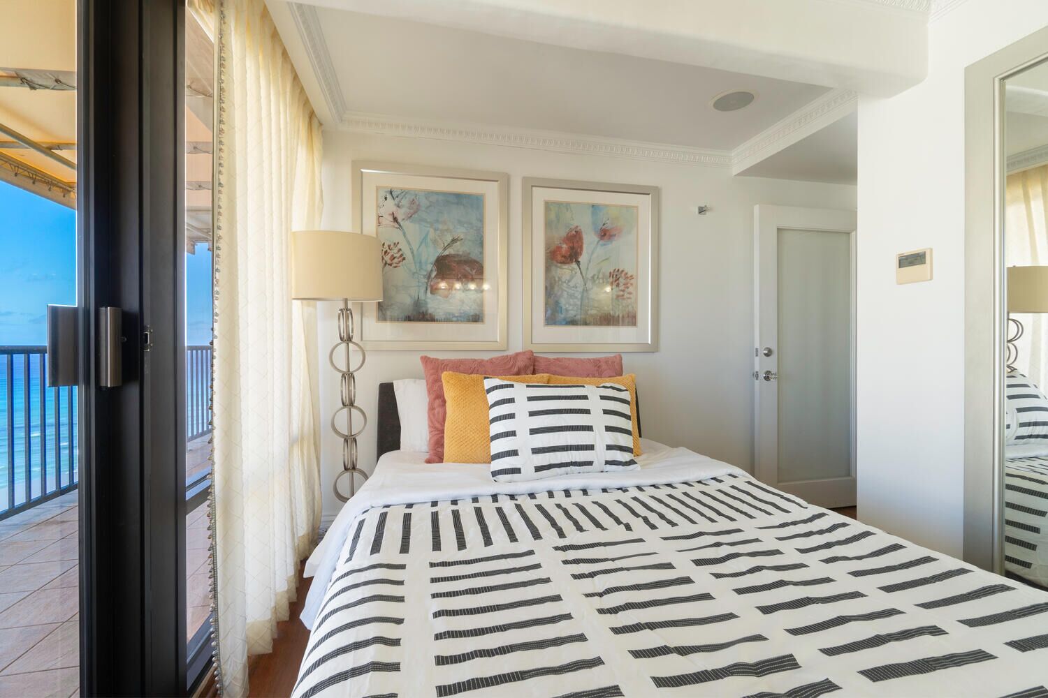 Une chambre d’hôtel avec un lit, deux tableaux accrochés au mur, une porte et une vue sur la mer depuis le balcon.