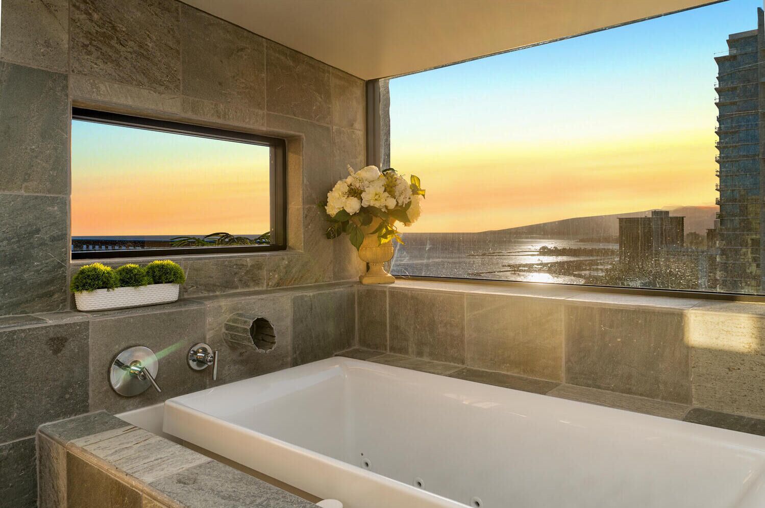 Ein modernes Badezimmer mit einer großzügigen Badewanne, einem Fenster mit Blick auf die Stadt und einer Vase mit Blumen.