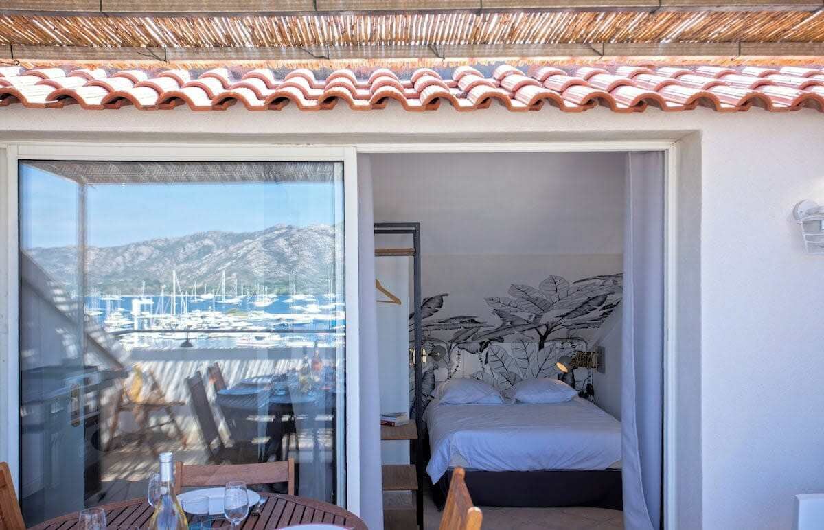 A room with a bed, a window with a view of boats, and a table with chairs.