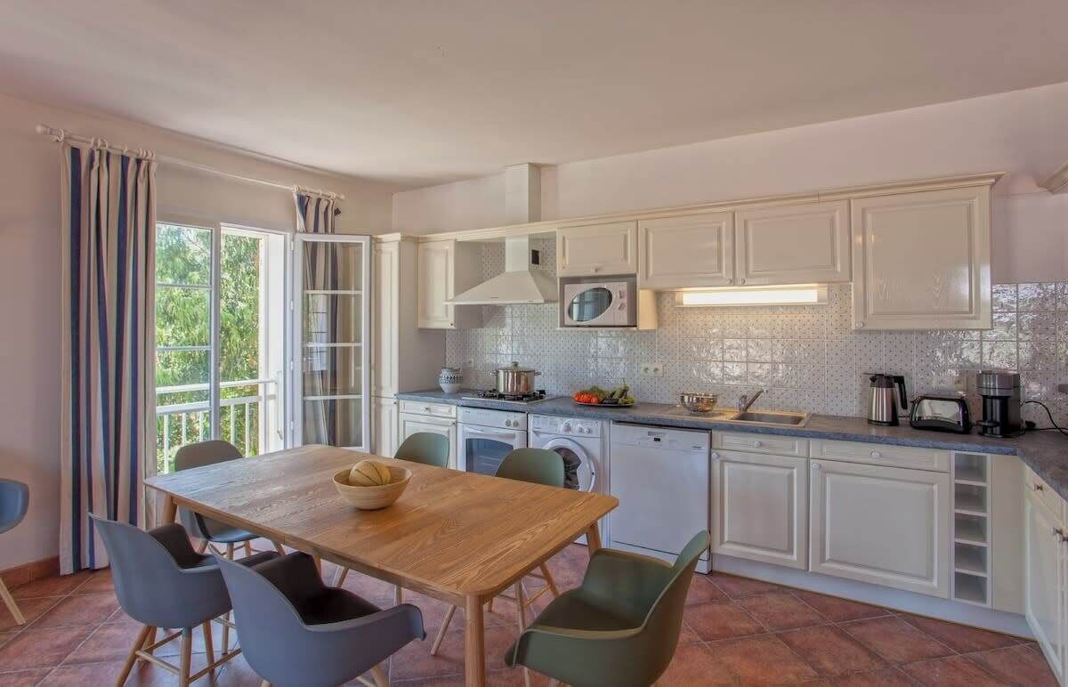 A modern kitchen with white cabinets, a wooden dining table, and a view of greenery outside.
