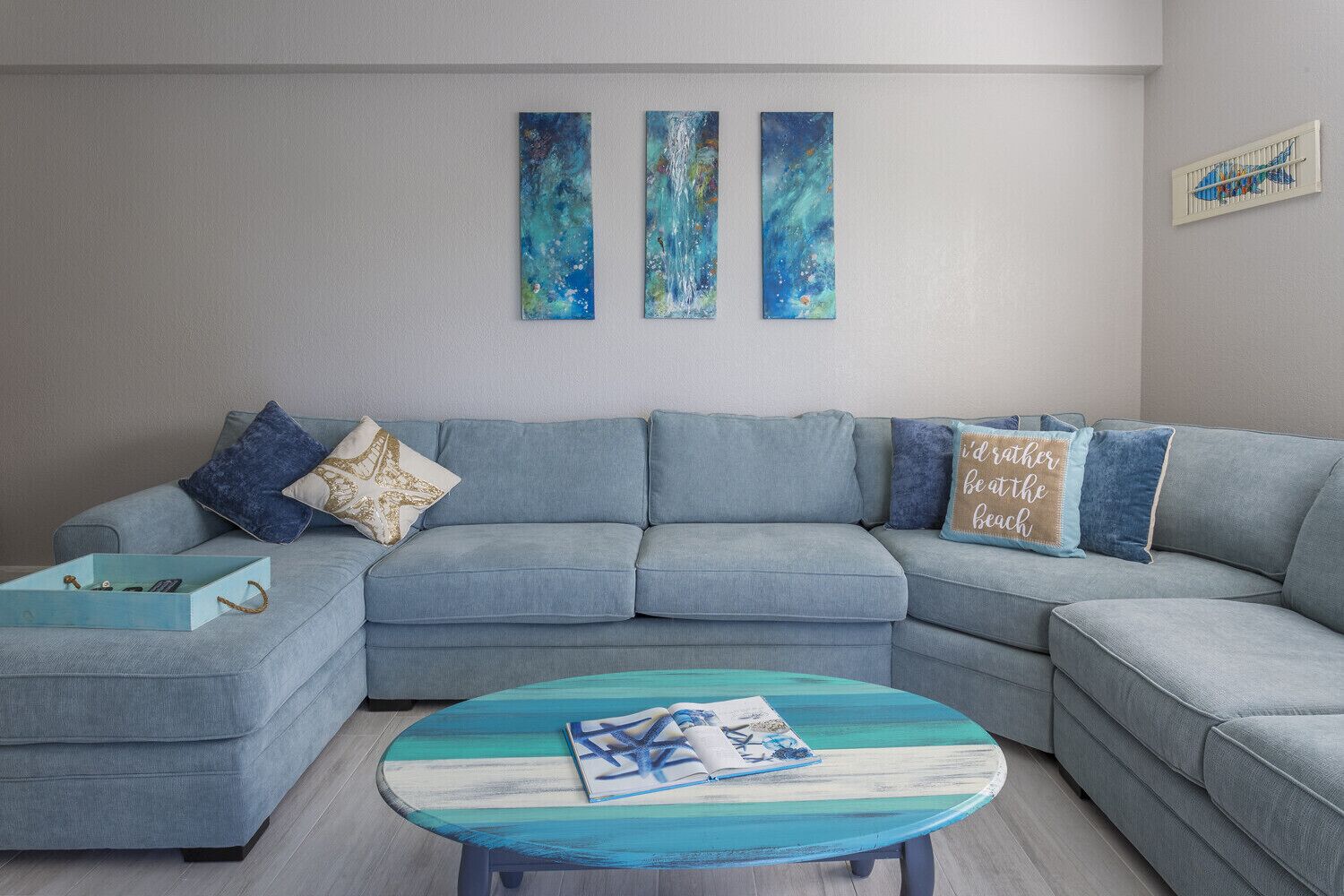 A modern living room with a grey sectional sofa, a round coffee table with a blue and white striped top, and three abstract paintings on the wall.