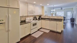Una cocina moderna con armarios blancos, microondas integrado, estufa y pileta. Hay dos alfombras en el piso y un ventilador de techo.
