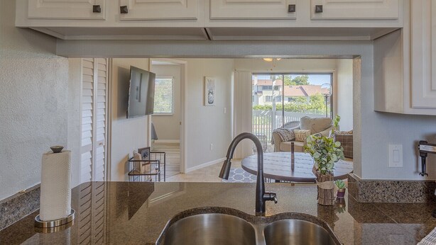 A modern kitchen with granite countertops, stainless steel sinks, and white cabinetry. There is a dining area with a round table and chairs, and a view of the outside through a sliding glass door. - La Casa Del Mar 608 Star5vacationscom (Dunedin)