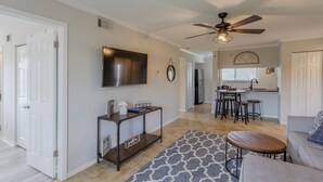 A spacious living room with a large sofa, a coffee table, and a patterned rug. There is a kitchen area with a dining table and bar stools. A ceiling fan and a mounted TV are visible. - La Casa Del Mar 604 Star5vacations (Dunedin)