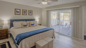 Quarto com cama, ventilador de teto, mesa de cabeceira, um banco e vista exterior através de uma porta de correr de vidro.