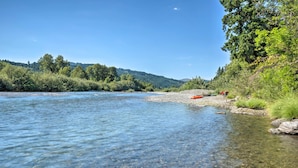 A clear river with a rocky shore and dense greenery in the background. - Quaint Riverside Reprieve w/ Kayaks + Floats! (Brookings)
