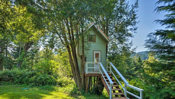 A treehouse with a wooden deck and a white railing, surrounded by lush greenery and trees. - Quaint Riverside Reprieve w/ Kayaks + Floats! (Brookings)