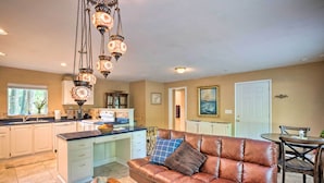 A spacious living area with a brown leather sofa, a wooden coffee table, and a dining table with chairs. A kitchen with white cabinets and a large island is visible in the background. - Quaint Riverside Reprieve w/ Kayaks + Floats! (Brookings)
