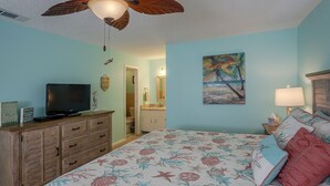 A bedroom with a bed, a television on a dresser, a ceiling fan, and a painting of a beach scene. - Beautiful Seashell/ Pool Star5vacations (Indian Shores)