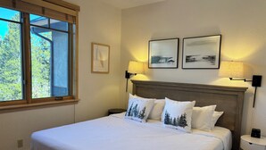 Una cama bien arreglada con sábanas blancas y almohadas con estampado de árboles, un cabecero de madera, dos lámparas de pared y una mesita de noche con una lámpara.