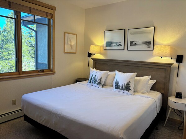 A neatly made bed with white linens and tree-patterned pillows, a wooden headboard, two wall-mounted lamps, and a small bedside table with a lamp.
