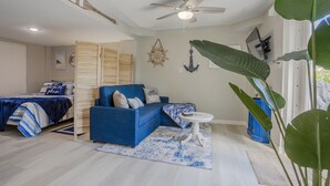 Ein Zimmer mit einem blauen Sofa, einem Bett mit blau-weiß gestreifter Bettdecke, einem Deckenventilator und maritimer Dekoration, darunter ein Schiffsrad und Anker-Wandbilder.