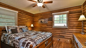 A log cabin bedroom with a wooden bed, a ceiling fan, a window with blinds, and a lamp on a dresser. - Rustic Cabin in the Woods Star5vacations (Black Mountain)