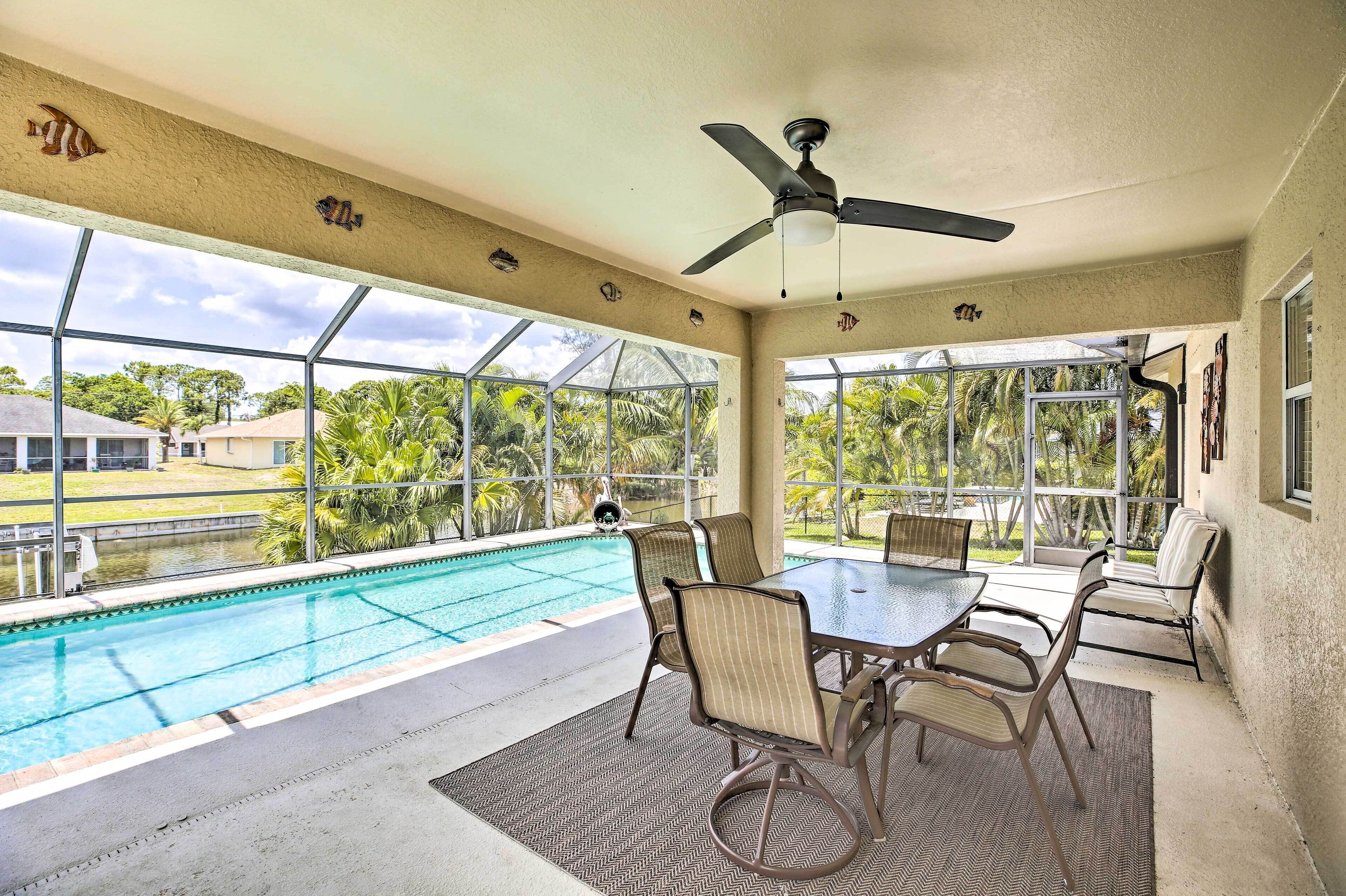 A poolside area with a table and chairs, a ceiling fan, and large windows offering a view of the outdoors.