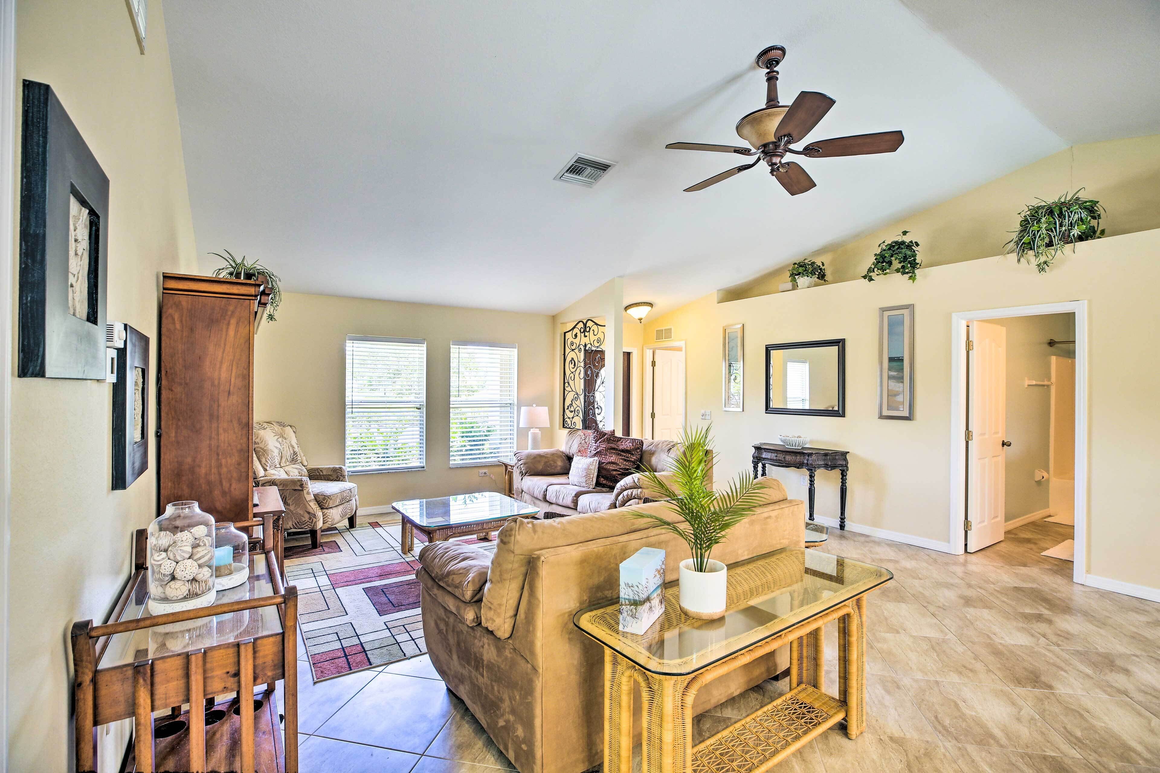 A spacious living room with a large sofa, a coffee table, and a dining table. There is a ceiling fan, a window with blinds, and a door leading to another room.