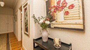 Un couloir avec un vase de fleurs posé sur un plateau noir, un tableau encadré accroché au mur et un luminaire au plafond.