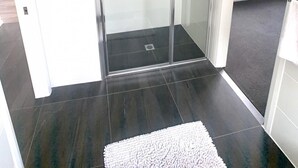 A modern bathroom with a glass shower enclosure, dark tiled flooring, and a white rug.