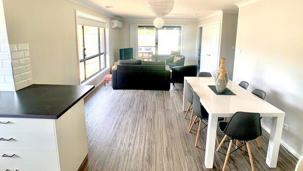 A modern kitchen with a black countertop and white cabinets, a dining area with a white table and black chairs, and a living area with a dark sofa and a hanging spherical light fixture.