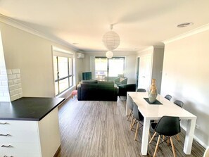 A modern kitchen with a black countertop and white cabinets, a dining area with a white table and black chairs, and a living area with a dark sofa and a hanging spherical light fixture.