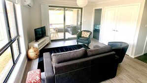 A modern living room with a sofa, a TV, and a chair. There is a large window and a door leading to a balcony.