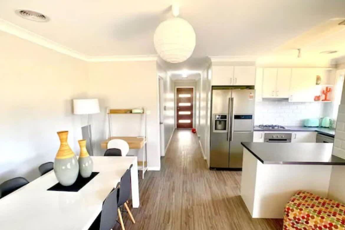 A modern kitchen with a central island, a dining area with a table and chairs, and a hallway leading to a bedroom.