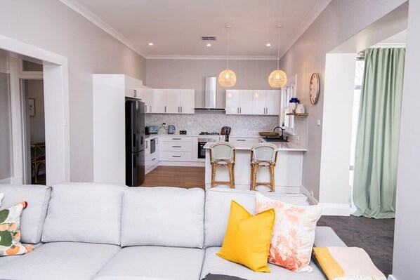 A modern kitchen with white cabinets, a dining area with wicker chairs, and a grey sofa with yellow and orange cushions.