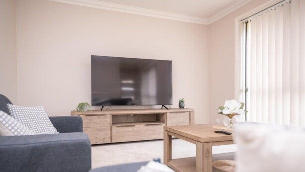 Uma sala de estar com televisão de tela plana em um suporte de madeira, um sofá cinza, uma mesa de centro de madeira e uma janela com persianas.