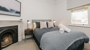 Een slaapkamer met een open haard, een bed met een grijs hoofdeinde, bedlampen en een raam met uitzicht.