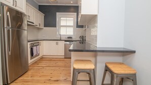 Una cocina moderna con armarios blancos, un refrigerador de acero inoxidable, horno empotrado y encimera negra. Hay dos taburetes de madera.