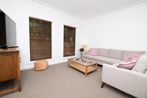 Una sala de estar con un sofá, una mesa de centro, un televisor sobre un mueble de madera y dos ventanas con persianas.