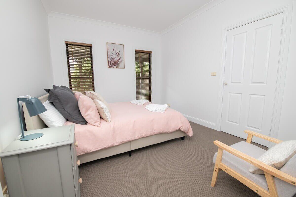 Un dormitorio con cama, mesita de noche, silla y dos ventanas.